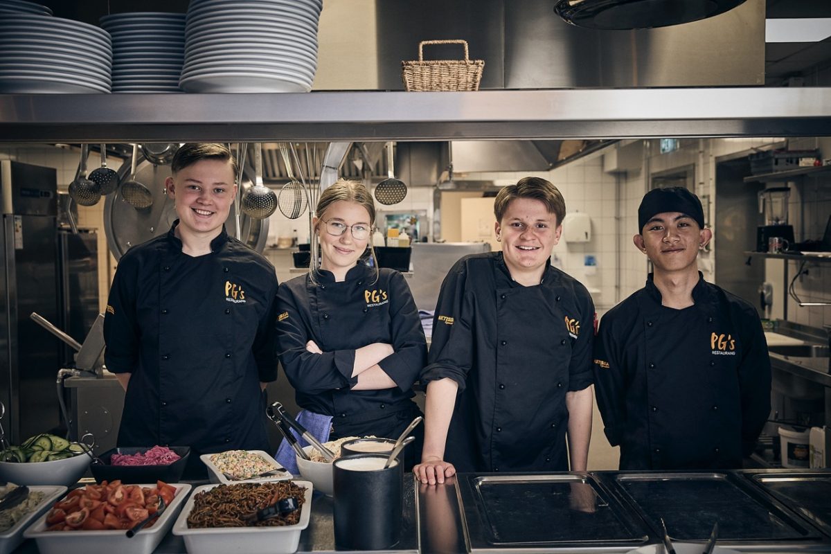 Fyra Praktiska elever i kockkläder i ett restaurangkök som ler mot kameran