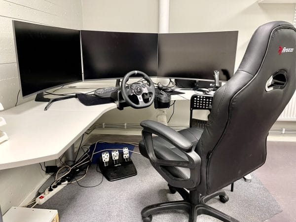 Skrivbord med tre datorskärmar, en datorstol och en ratt.