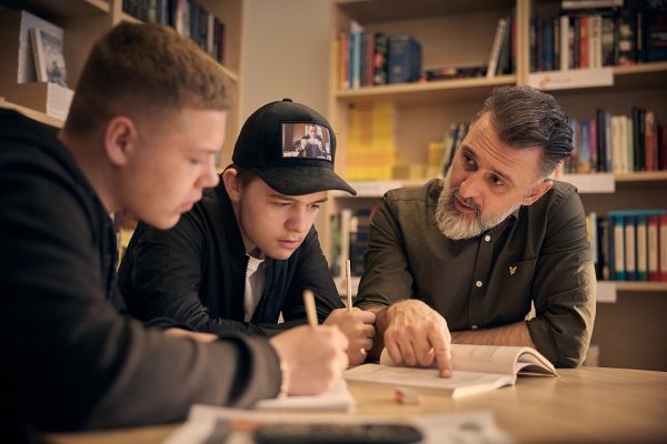 Två elever som studerar i biblioteket tillsammans med en lärare.